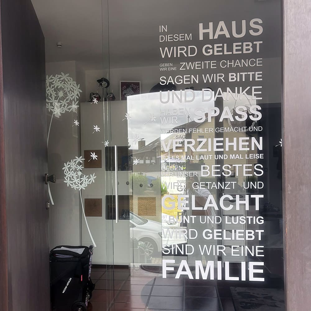 Weißer Wandtattoo-Spruch In diesem Haus auf einer Glastür mit zusätzlicher Pusteblumen-Dekoration